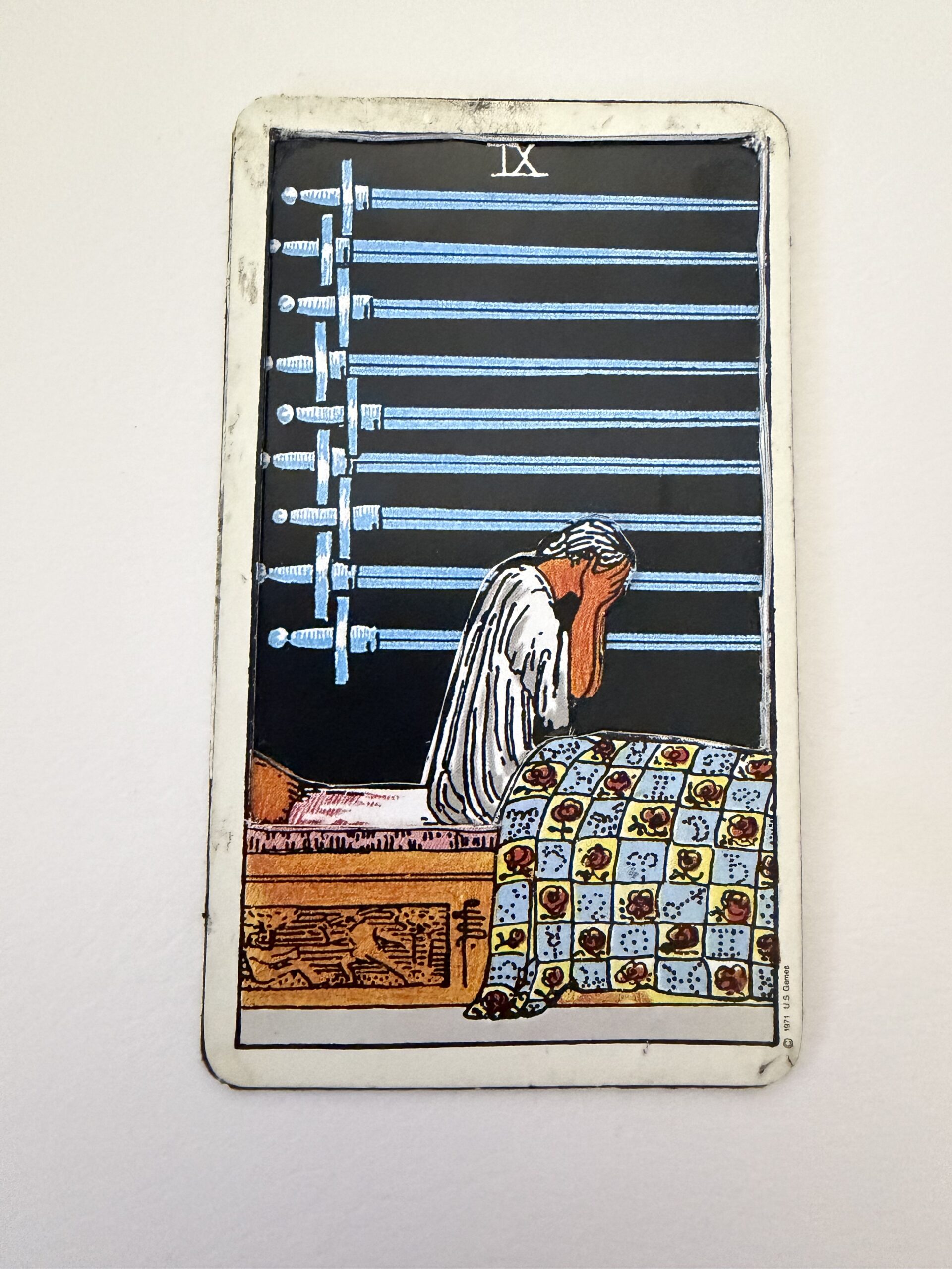Nine of Swords tarot card showing figure with head in hands sitting on bed with patterned quilt, nine swords mounted on black wall above, vintage Rider-Waite deck